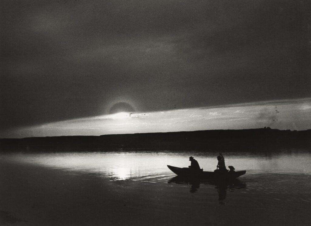 Fotografía en blanco y negro. Dos hombres atravesando el río con una pequeña barca.
