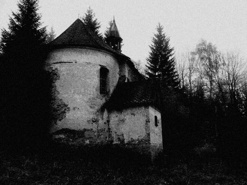 La imagen es una fotografía en blanco y negro de una pequeña capilla circular en medio del bosque.
