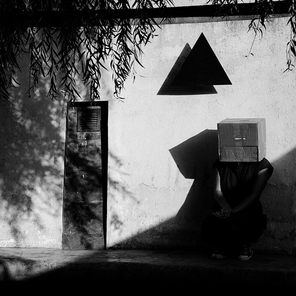 La foto muestra a un hombre en cuclillas apoyado en la pared de una pequeña caseta.. El hombre tiene la cabeza tapada con una caja de cartón. A su derecha hay una puerta estrecha. Arriba de él, dos triángulos negros superpuestos.
